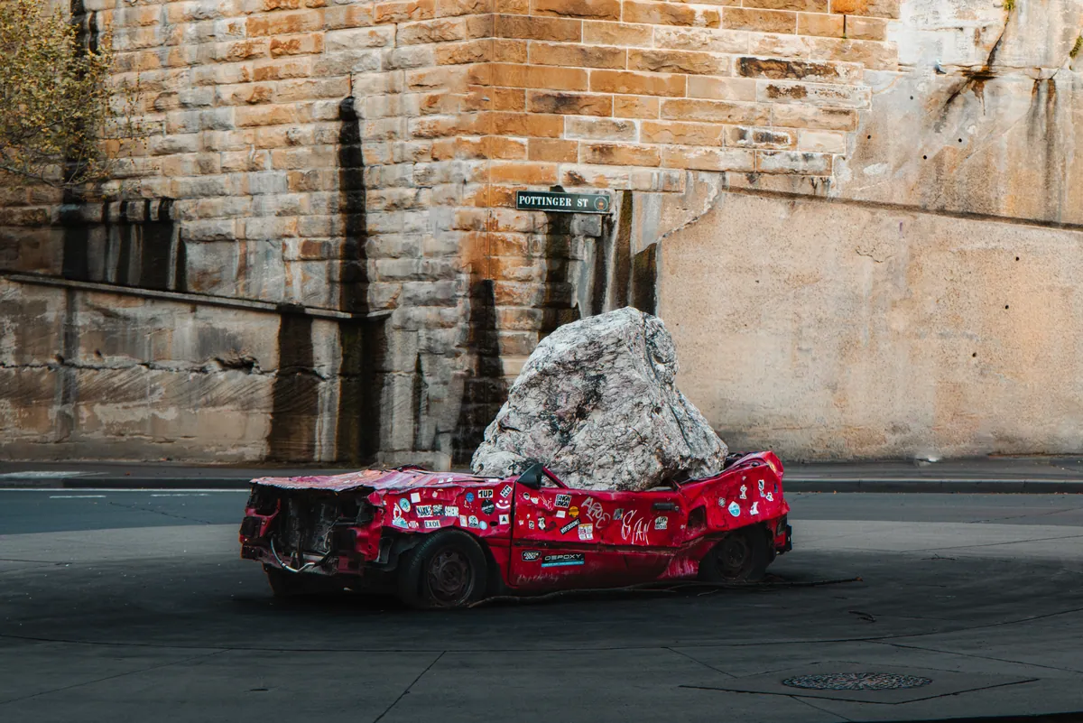 A crushed red car pinned under a boulder, a sculpture against sandstone walls on Pottinger Street