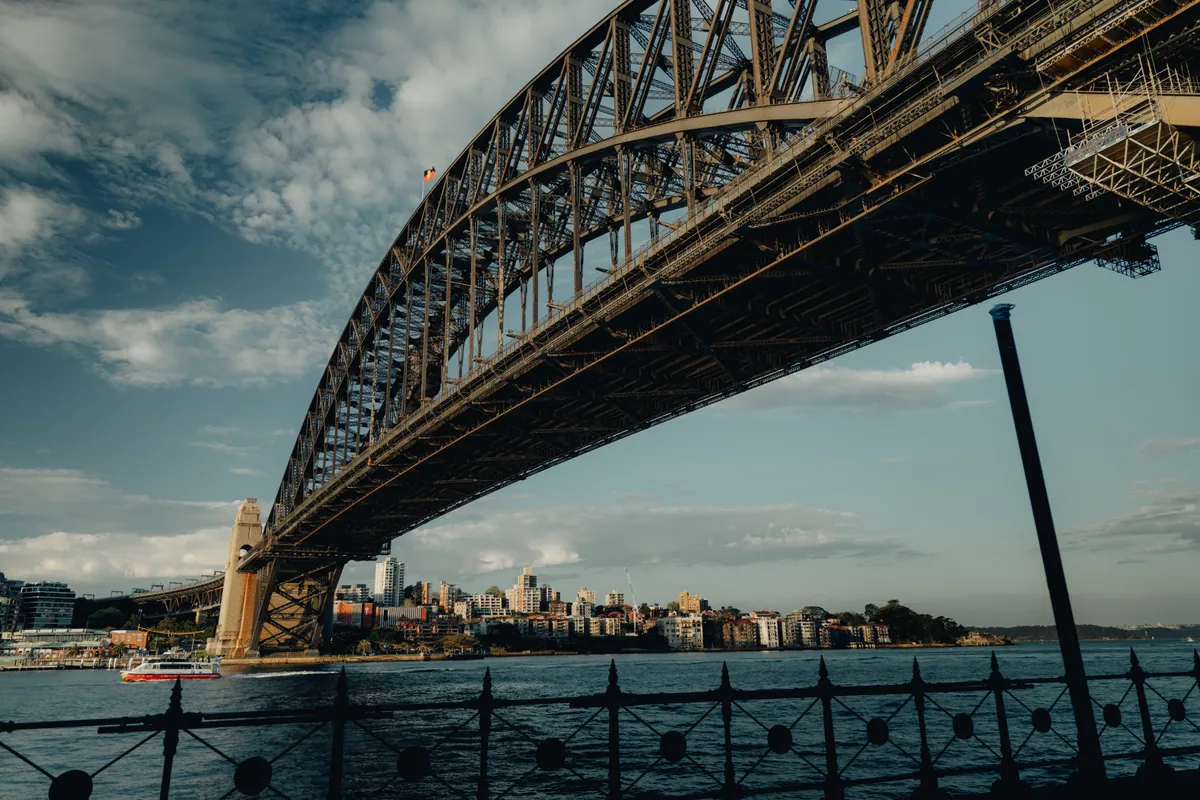 The Sydney Harbour Bridge seen from below, its steel arch crossing a cloud-streaked sky